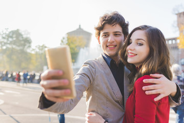 Young beautiful couple in love taking selfie outdoor in the city with smart phone hand hold - vanity, sharing, social network concept