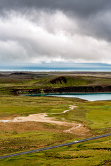Fototapeta na wymiar Icelandic landscape with blurry moving car, black road, green scenery, a blue water lake and spectacular cloudy sky