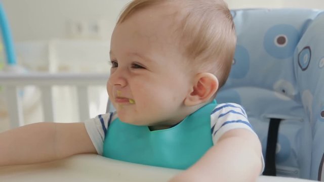 Closeup of young mother feeding cute smiling baby boy wearing bib and sitting in highchair