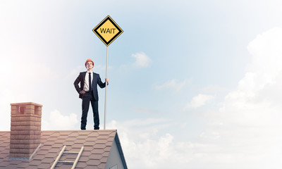 Young businessman on house brick roof holding yellow signboard. 