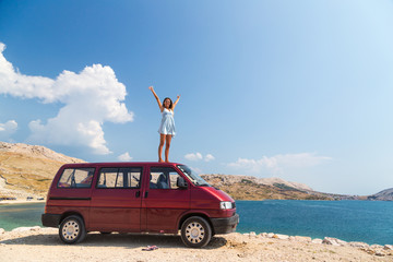 Beautiful tanned girl in a blue dress standing on a rooftop of red van and spreading arms. © paul prescott
