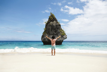 Back view of young hipster standing barefooted on sandy coastline, facing rocky cliff in the middle...
