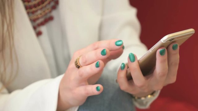 Hands of young woman in white jacket holding and using gold smartphone. Close-up