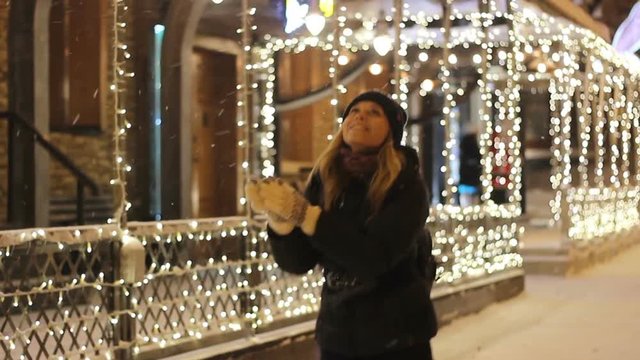 Young happy woman catches snowflakes on the background of lights in winter night