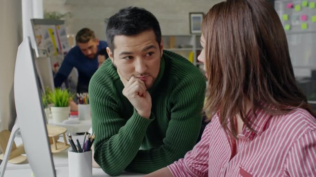 Laughing Asian man standing near cheerful female colleague talking on mobile phone, then listening to her and smiling as she showing him something on computer in office 