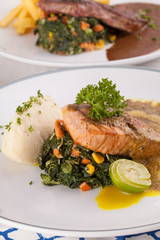 A decorating nice dish of salmon steak and beef served with mashed potato,lemon, saute spinach and sauce on background with wooden utensils.