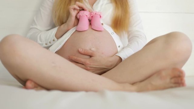 Waiting for baby with baby's booties. Young pregnant woman sits and stroking belly. She holds the pink baby's bootees.