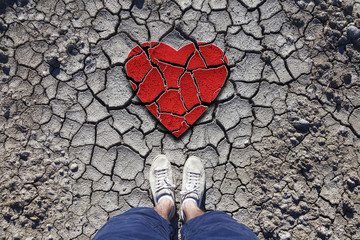 Lonely man standing on dried soil ground with illustrated cracked heart symbol. Conceptual broken love photo. Point of view perspective used.
