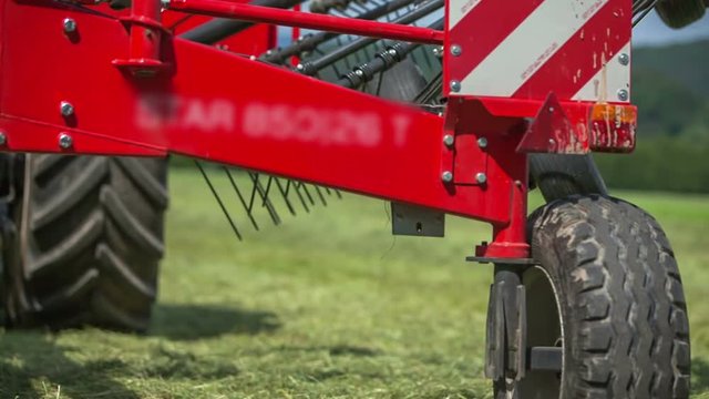 A red trailor is standing still and rotary rakes are moving. A farmer will now start preparing hay on a big field.
