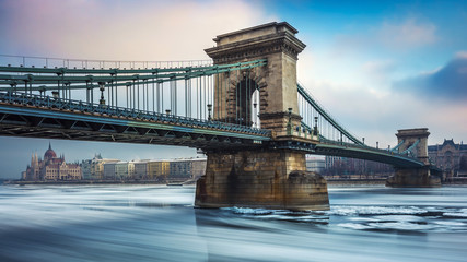 Fototapeta premium Budapest, Hungary - The iconic Szechenyi Chain Bridge on the icy River Danube on a cold winter morning with Hungarian Parliament at the background
