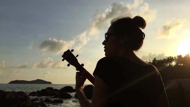 Young mixed race girl playing the ukulele on the beach rocks. Colorful sunset. HD back view silhuette. Thailand.