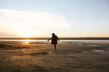 Naklejka premium Beautiful girl is standing near sea on winter and see to the sundown, black coat, beautiful eyes, model, a beautiful sunset, the sun's rays, reflection in water