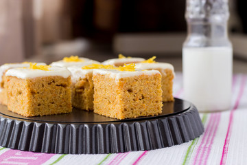 Pieces of homemade carrot cake with orange, zest and icing cream, milk in small bottle, on the dark background. Selective focus