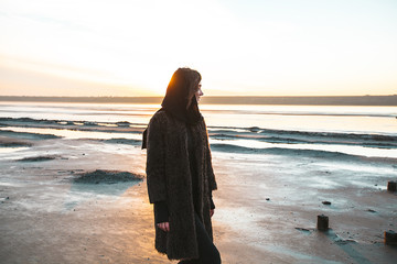 Beautiful girl is standing on the sea in a Sunbeam, yellow boots, long black coat, sand underfoot