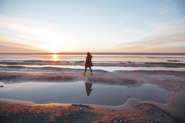 Beautiful girl is standing on the sea in a Sunbeam, yellow boots, long black coat, sand underfoot