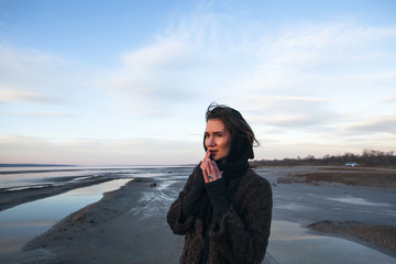Beautiful girl is standing on the sea in a Sunbeam, yellow boots, long black coat, sand underfoot
