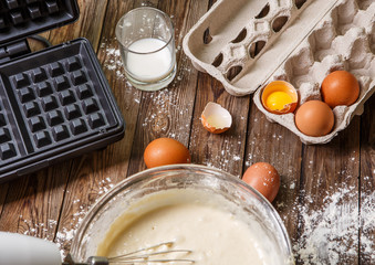 Making waffles at home - waffle iron, batter in bowl and ingredients - milk, eggs and flour.