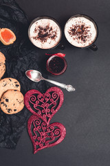 Romantic breakfast. Two Cups of coffee, cappuccino with chocolate cookies and biscuits  near red hearts on black table background. Valentine day. Love. Top view. 