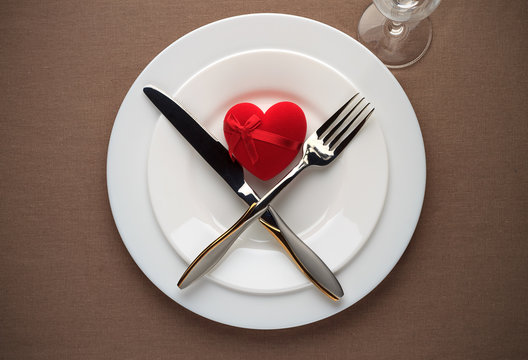 Valentines day table setting with red heart, white plates, fork, knife and wineglass.