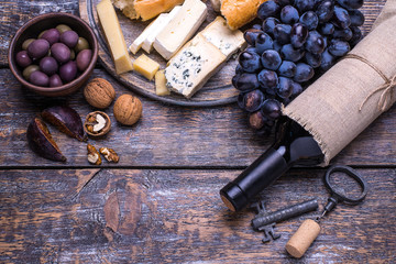 Red wine in a bottle, cork, bottle screw and a set of products - cheese, grapes, nuts, olives, figs on a wooden board, background