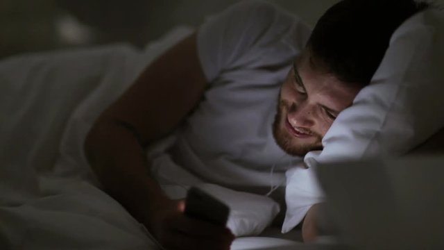 man with smartphone and earphones in bed at night