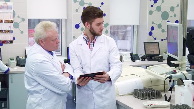 Brunette young man showing something on monitor to his colleague at the laboratory. Gray senior scientist looking at what his intern demonstrating. Brunette lab worker holding tablet in his hands