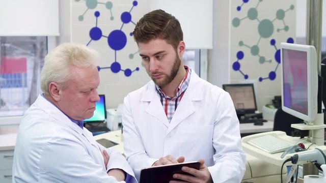 Attractive male scientist showing something on his tablet to his coworker at the laboratory. Two caucasian men in white coats looking at the tablet screen. Gray senior man holding his hands crossed on