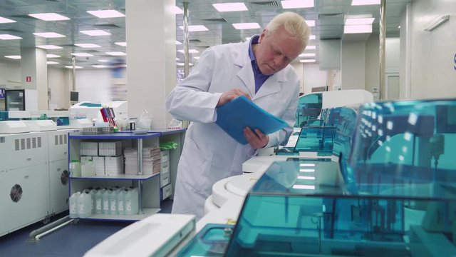  Gray aged man standing near laboratory devices. Senior lab worker looking at scientific equipment. Mature male scientist standing on background of working centrifugal machine