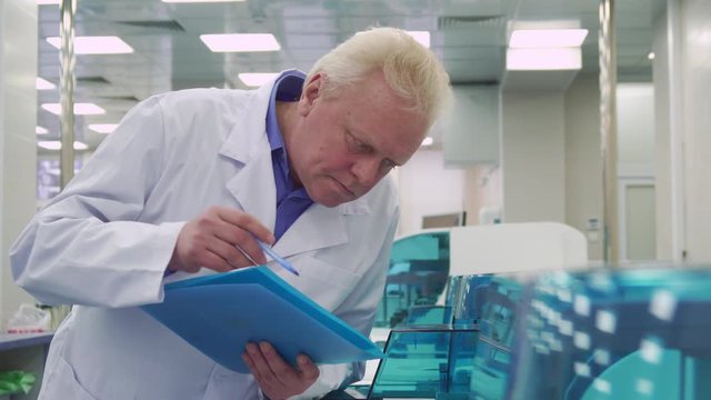Mature caucasian man in white coat noting report about laboratory machine work. Senior male scientist looking at his list with data. Gray lab worker tilting above the centrifugal machine