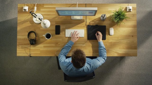 Top View of a Young Graphic Designer Working on a Digital Drawing Tablet Sitting at His Table. Also there is Desktop Computer, Headphones and Smartphone on the Table. 