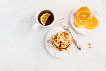 Stack of homemade pancakes with banana, maple syrup and walnuts on vintage plate. Fork, fresh sliced fresh lemon, cup of tea, white and gray concrete background, top view.