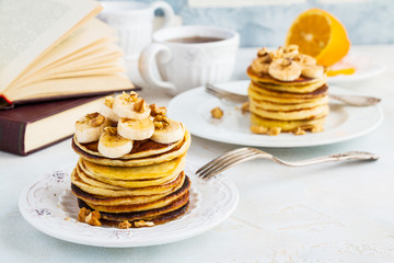 Stack of homemade pancakes with banana, maple syrup and walnuts on vintage plate. Fork, fresh sliced fresh lemon, cup of tea, open book, white and gray concrete background.