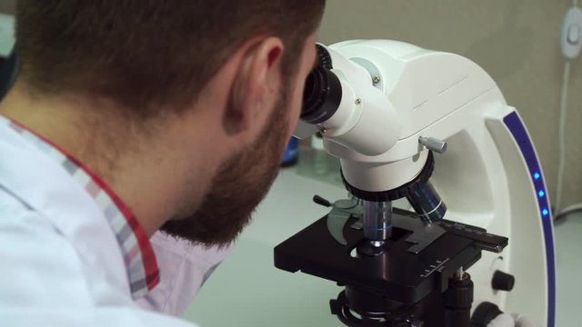 Young brunette man spinning coarse adjustment knob. Close up of attractive bearded male scientist zooming image on the scope. Young lab worker moving the part of microscope at the laboratory