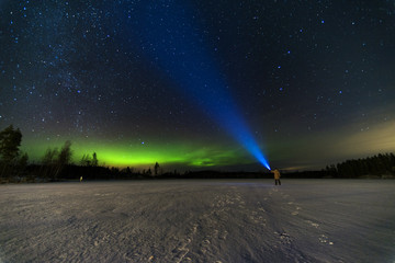 person puts bright blue flashlight to the starry sky with green north lights using torch staying on frozen lake 