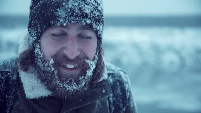 Young man with beard and long brown hair with snowbound hat, jacket and face smiling at camera
