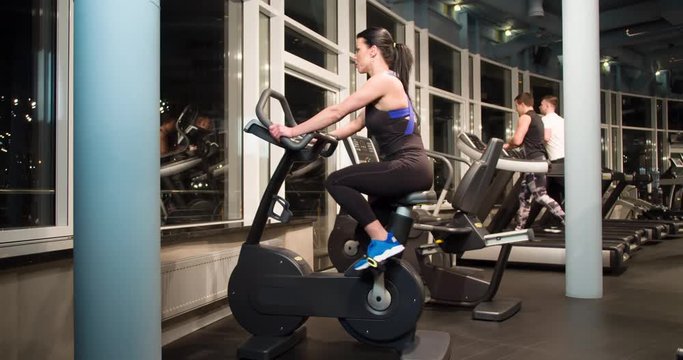 Woman on cardio training in gym 4k close-up video. Fitness girl working out with stationary bicycle. Male athletes running at on background 