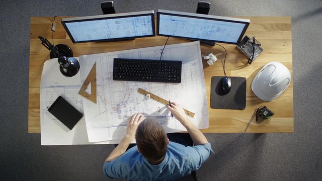 Top View of a Technical Engineer Working on His Blueprints, Drawing Plans, Using Desktop Computer. Various Useful Items Lying on his Table.  Shot on RED Cinema Camera in 4K (UHD).