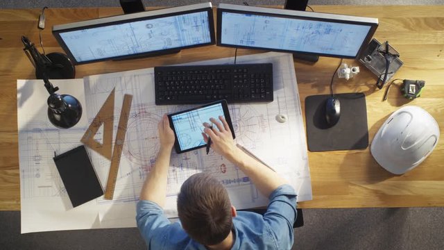 Top View of architectural Engineer Draws on His Blueprints, Compares with Tablet Computer with Green Screen, Using Desktop Computer Also. His Desk is Full of Useful Objects and Evening Sun. 