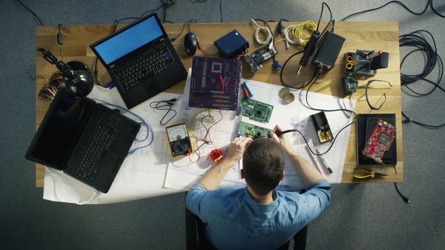 Top View of Young IT Technician Soldering Circuit Board. His Table is Full With Various Computer Parts, Motherboards, Laptops, Cables. Morning Sun Illuminates His Desk.