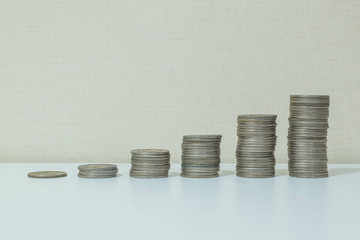 Closeup pile of coin start from low to high on white wood desk and cream wallpaper textured background with copy space