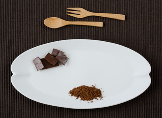Flat lay image of rectangle dark chocolate pieces and 100% cocoa powder on ceramic white plate shaped as banana leave over dark brown cotton place mat with wooden spoon and fork