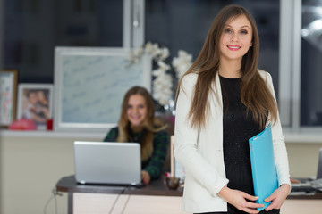 Beautiful smiling businesswoman holding file in office and woman working in the background.