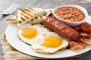 Traditional full English breakfast with fried eggs, sausages, beans, mushrooms, grilled tomatoes and bacon on wooden background
