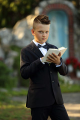 Pierwsza komunia, chłopiec w garniturze z książeczką na tle kapliczki.
