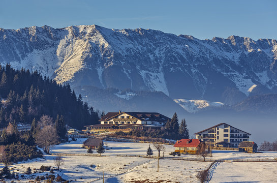 Sunny winter scenery with majestic snowy Piatra Craiului mountains, chalet or lodging buildings in Cheile Gradistei Fundata touristic resort, Romania. Idyllic, quiet holiday retreat destinations.