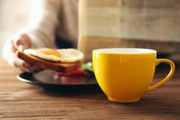 Cup of tea and tasty breakfast on table with person reading newspaper on background
