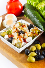 Simple, fresh salad with vegetables and cheese in white bowl on a wooden board next to cucumber, tomato, olives and egg.