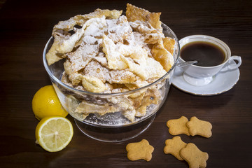 Sprinkle with powdered sugar, fried plate of shortcake dough.Dessert Polish cuisine - faworki(angel wings). Still life. Faworki in a glass vase,coffee in a porcelain cup,lemons and shortbread .