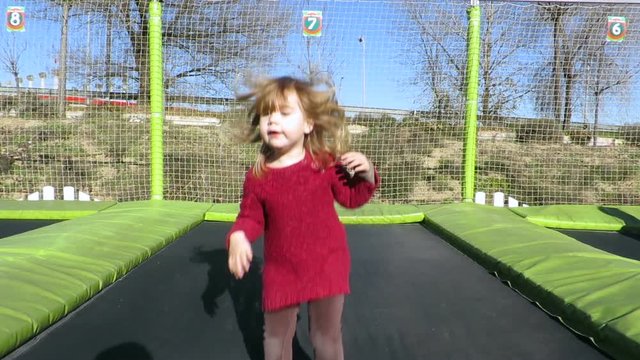 slow motion 4k video. happy three years old blonde child with winter red sweater trampolining or jumping on trampoline in outdoor playground
