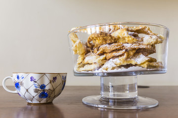Sprinkle with powdered sugar, fried plate of shortcake dough. Dessert Polish and Lithuanian cuisine - faworki ( angel wings). Faworki in a large glass vase on a beige background.